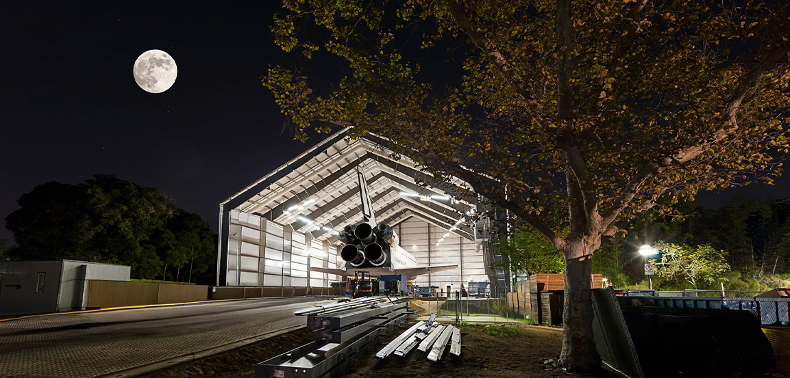 Space Shuttle moving into large Butler Building hangar under a full moon.